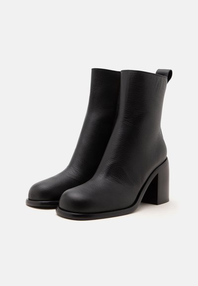 Paire de bottines en cuir noir avec bouts arrondis, talons blocs et languette à l’arrière, sur un fond clair uni.