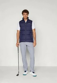 Σκούρο μπλε γιλέκο με φερμουάρ, φορεμένο πάνω από ένα άσπρο πουκάμισο. Ανοιχτό γκρι παντελόνι γκολφ και λευκά παπούτσια με μπλε λεπτομέρειες. Ράκετ γκολφ κρατημένη στο δεξί χέρι.