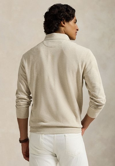 Beige polotrøye med lange ermer, med krage, ribbestrikkede mansjetter og teksturert stoff. Brukt med hvite bukser, sett bakfra.