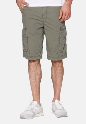Cargo-Shorts aus olivgrünen Stoff, mit Seitentaschen, ausgefransten Kanten und kontrastierendem Nähen. Kombiniert mit hellen Sneakers.