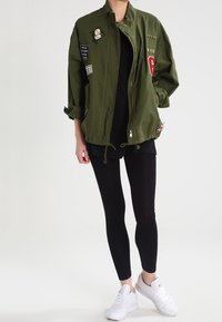 Olchowa, oversize'owa kurtka z różnymi naszywkami, złotymi akcentami i zamek błyskawiczny. Czarny top i legginsy, w połączeniu z białymi sneakami.