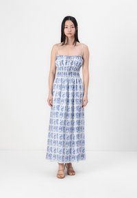 Abito maxi in tessuto leggero, con un motivo floreale blu e bianco, corpino smockato e spalline sottili regolabili. Sandali marroni completano il look.