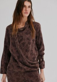 Haut à motif floral marron avec un devant drapé, des manches longues et des accents dorés au niveau des poignets. Tissu doux avec une texture lisse.
