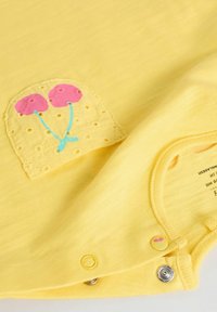 Un body en coton jaune avec un patch orné de dentelle, décoré de ballons roses et de cordons bleus. Les fermetures à pression en bas augmentent la praticité.
