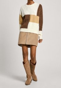 Pull en maille avec un motif patchwork en crème, beige et marron. Assorti à une jupe en velours côtelé beige et des bottes cowboy en daim marron.