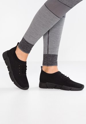 Czarne dziergane sneakersy na sznurówki noszone z szaro-białymi paskowanymi legginsami na białej podłodze, pokazujące widok z boku i z tyłu.