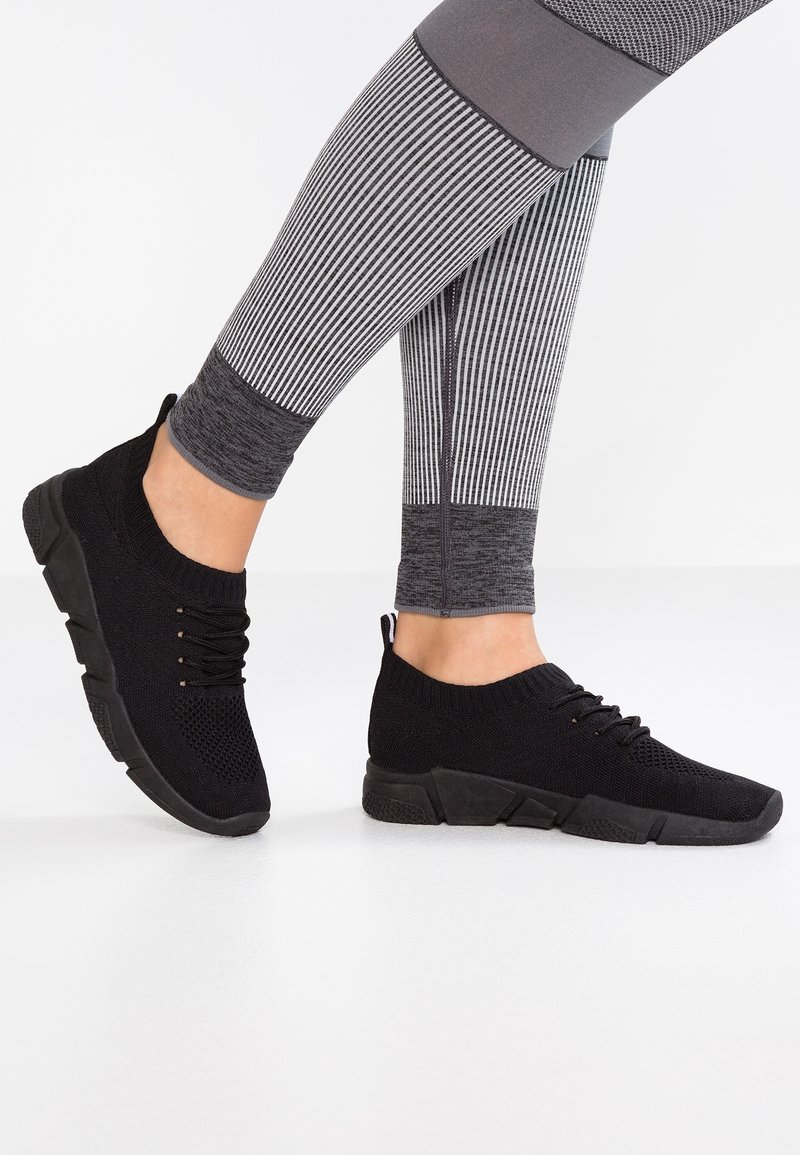 Baskets en maille noires à lacets portées avec des leggings à rayures grises et blanches sur un sol blanc, montrant les vues latérales et arrière.