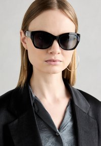 Schwarze übergroße Sonnenbrille mit glänzender Oberfläche, kantiger Form und dunklen Gläsern. Getragen mit einem schwarzen Blazer und einem grauen Satink Hemd.