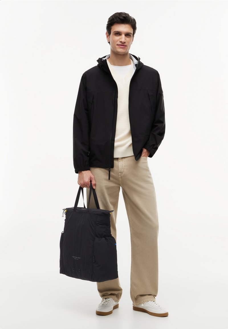 Jeune homme portant une veste à capuche noire, un pull crème, un pantalon beige, des baskets blanches, tenant un grand sac fourre-tout noir avec deux poignées.
