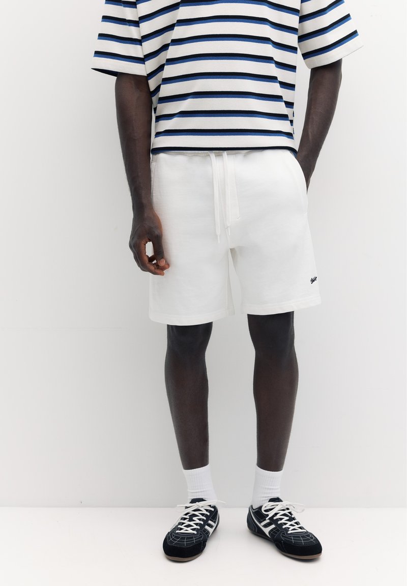 Personne portant un short blanc à cordon, une chemise rayée bleue et blanche, des chaussettes blanches et des baskets noires à lacets, debout devant un fond blanc.