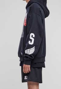 Sudadera con capucha negra que presenta un estampado gráfico rojo en la parte frontal, detalles de logo blancos en las mangas, y combinada con pantalones cortos de malla negros.