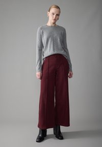 Mujer vestida con un suéter gris de manga larga, pantalones burdeos de pierna ancha de cintura alta, botas negras de tacón y pendientes de aro plateados, de pie.