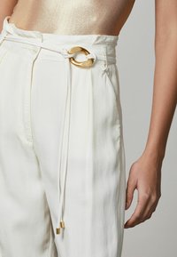 Pantalones blancos de cintura alta cuentan con un hebilla decorativa en tono dorado y un cierre con cordón, elaborados con una tela suave y ligera.
