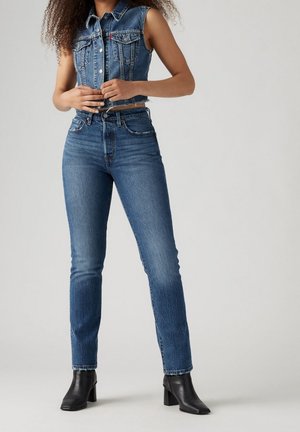 Vrouw die een blauw hoog getailleerde spijkerbroek, een cropped mouwloos spijkerjasje en zwarte enkellaarsjes met hak draagt, staand tegen een effen achtergrond.