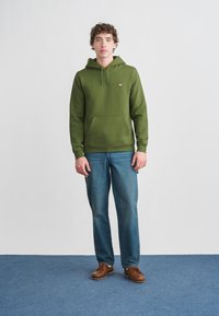 Φόρμα με κουκούλα σε ελιά πράσινο χρώμα με τσέπη καγκουρό και κορδόνι στην κουκούλα, σε συνδυασμό με ανοιχτόγαλα τζιν και καφέ δερμάτινα παπούτσια με κορδόνια, πάνω σε μπλε χαλί.