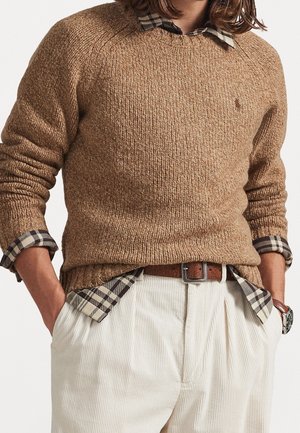 Homme portant un pull en maille marron par-dessus une chemise à carreaux aux manches retroussées, un pantalon en velours côtelé blanc, une ceinture marron et une montre-bracelet.