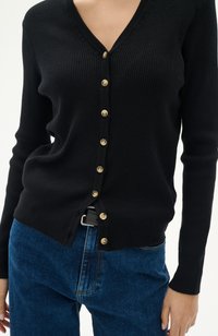 Svart ribbad cardigan med V-ringning, prydd med guldknappar framtill. Bärs över blå denimjeans, vilket framhäver kontrasterande färger och texturer.