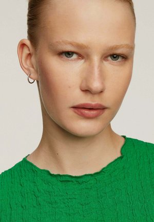 Top verde texturizado con escote en punta, combinado con un pequeño arete de plata en forma de aro. Presenta un maquillaje natural y un fondo neutro.