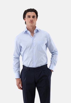 Helles blau gestreiftes Hemd mit Knopfverschluss und einer einzelnen Brusttasche, kombiniert mit dunklen marineblauen Hosen, die Falten und seitliche Taschen haben.