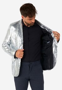 Silver sequin blazer with a shiny surface, notched lapels, and black satin lining shown open. Black shirt and dark patterned trousers worn underneath.