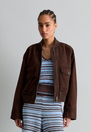 Junge Frau mit geflochtenem Haar, die eine braune Jacke mit Reißverschluss über einem blau-braun gestreiften gestrickten Top und passendem Rock trägt, steht vor grauem Hintergrund.