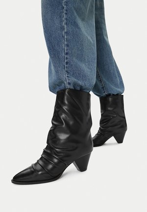 Schwarze, lockere Stiefel aus Leder, die bis zur Mitte der Wade reichen, mit spitzen Zehen und niedrigen Kegelhöhen, getragen zu lockeren blauen Jeans.