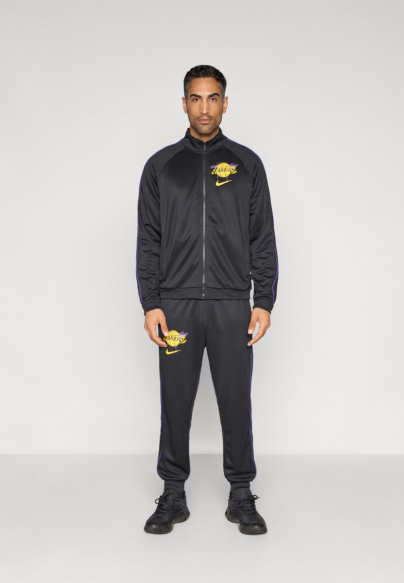 Μαύρο σετ φόρμας Nike με ζακέτα με φερμουάρ, μοβ ρίγες και λογότυπο Lakers, συνδυασμένο με αντίστοιχο παντελόνι και μαύρα αθλητικά παπούτσια.
