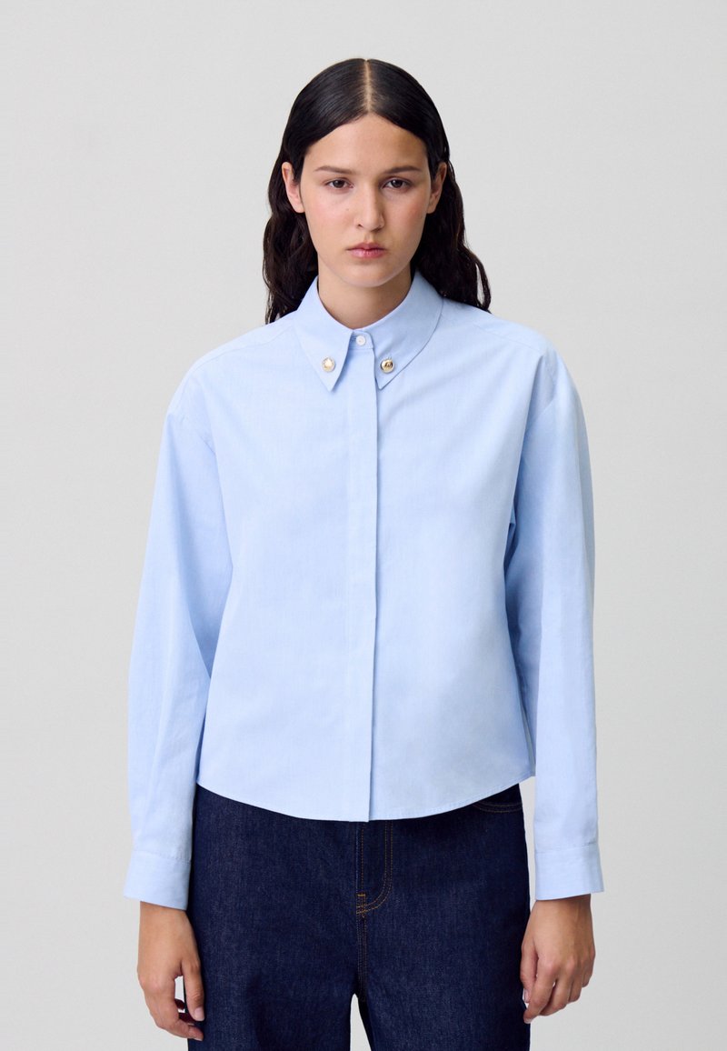 Chemise bleue clair à boutons avec un col arrondi, des manches longues et deux boutons dorés sur le col. Tissu doux avec une texture lisse.