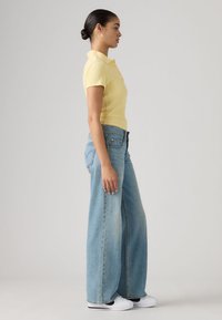 Κίτρινη ριγέ μπλούζα πόλο, ανοιχτό μπλε τζιν παντελόνι με ψηλή μέση και φαρδιά πόδια, και λευκά αθλητικά παπούτσια, ορατά από το πλάι με ουδέτερο φόντο.