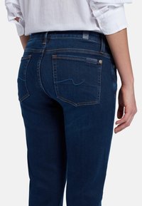 Jeans en denim bleu foncé avec une coupe classique. Présentent des poches arrière avec des coutures discrètes, une étiquette sur la ceinture, et une texture lisse.