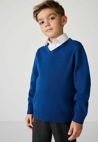 V-hals trui van effen blauwe gebreide stof, met ribgebreide manchetten en zoom. Gedragen over een witte blouse met kraag, gecombineerd met een donkere broek.