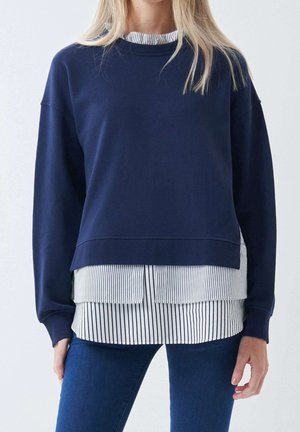 Femme portant un sweat-shirt bleu marine superposé à une chemise blanche à boutons avec des rayures verticales noires, associée à un jean bleu foncé.