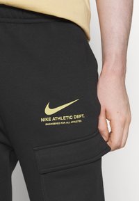Svarta träningsbyxor med en gul Nike-logotyp och text på låret, tillverkade av mjukt material med detaljer i form av en cargoficka.