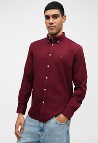 Bordo košulja s dugim rukavima i ovratnikom, koja sadrži malu logotip detalj, uparen s svijetloplavim trapericama. Glatka tekstura.