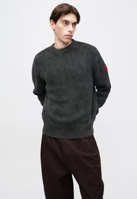 Tumši pelēks adīts džemperis ar raksturīgu riboto tekstūru, apaļu apkakli un sarkanu uzliktu plāksteri uz kreisā piedurknes.