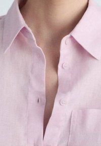 Helles Rosa, knöpfbares Hemd aus einem weichen, strukturierten Stoff. Verfügt über einen Kragen und eine kleine Brusttasche auf der linken Seite.
