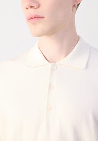 Polo shirt de couleur crème avec un tricot texturé, présentant un col classique, une patte de boutonnage à trois boutons et un léger éclat dans le tissu.