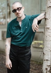 Polo verde de manga corta con cuello y botonera, combinado con pantalones negros. Modelo usando gafas de sol oscuras, de pie junto a un árbol.
