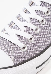 Baskets en maille grise avec lacets blancs, semelle noire contrastante et œillets circulaires. Surface texturée avec un motif en losange.