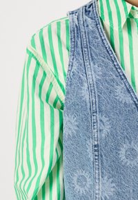 Light denim vest with floral pattern, worn over a green and white striped shirt. Visible textures and stitching details.