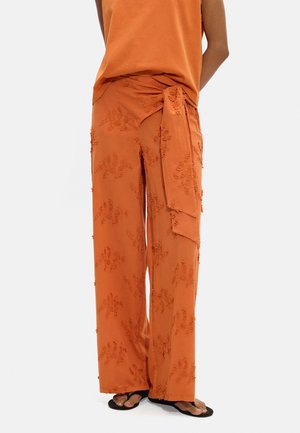 Spodnie o szerokich nogawkach w kolorze rdzy z fakturowanym wzorem kwiatowym i pasującym paskiem zawiązanym w talii, noszone z czarnymi sandałami.
