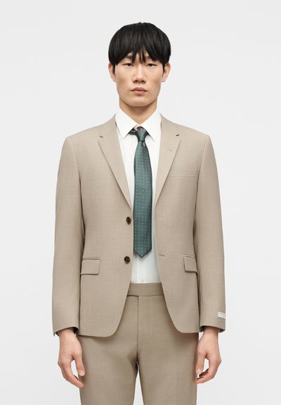 Μπεζ σακάκι κοστουμιού με δύο κουμπιά, ταιριαστό παντελόνι και ανοιχτό μπλε πουκάμισο. Δεμένη πράσινη γραβάτα με λευκές τελείες· υφασμάτινη υφή απαλή.