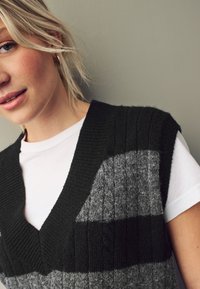 Strikket vest i mørkegrønne og grå nuancer, med en dyb V-hals og forskellige ribbede teksturer. Bæres over en hvid T-shirt.