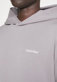 Sudadera con capucha gris de tela suave, que presenta un logo bordado en blanco de "Calvin Klein" en el pecho y un corte relajado.