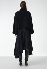 Zwarte cropped jack met een hoge kraag, gecombineerd met een gelaagde zwarte rok met streepjes en kniehoge zwarte laarzen.