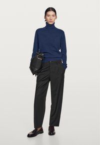 Marineblauwe coltrui, zwarte op maat gemaakte broek en bordeauxrode instappers. Model houdt een zwarte tas vast; de outfit heeft strakke lijnen en gestructureerde materialen.