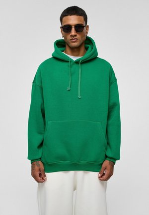 Groene oversized hoodie van een katoenmix, met een voorkantzak, trekkoordcapuchon en ribbelmanchetten. Draagt over een wit shirt.
