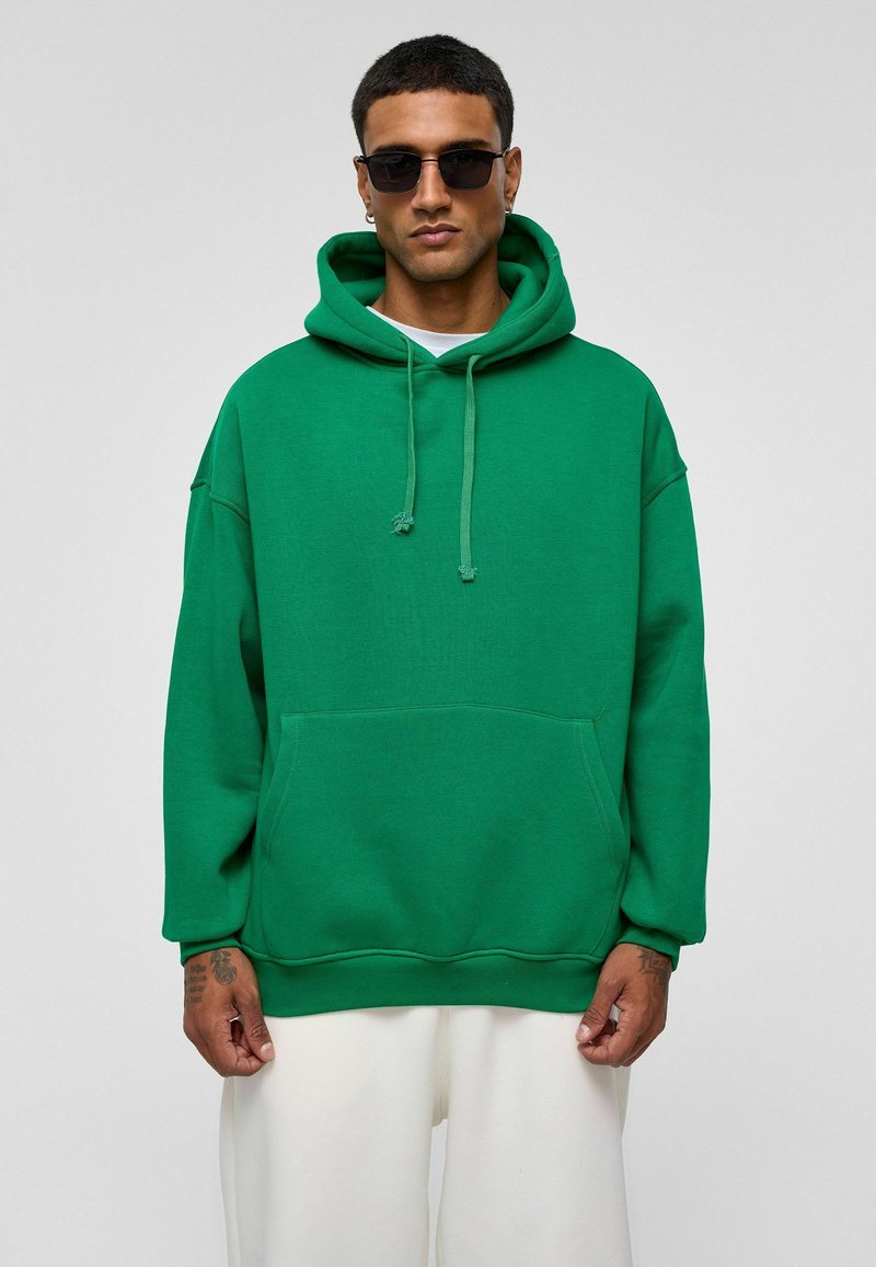 Groene oversized hoodie van een katoenmix, met een voorkantzak, trekkoordcapuchon en ribbelmanchetten. Draagt over een wit shirt.