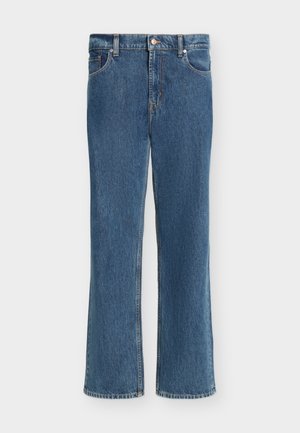 Jeune homme portant un cardigan beige sur une chemise à carreaux marron, un jean bleu et des chaussures marron foncé, debout avec une main dans la poche.