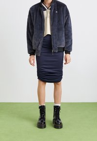 Zwarte nepbont bomberjack, beige crop top, marineblauwe gerimpelde pantalonrok en zwarte veterschoenen met witte sokken, neergelegd op een groene vloerbedekking.